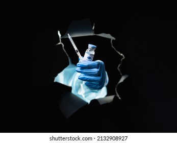 Vaccine Shot. Covid-19 Jab. Disease Prevention. Doctor Hand In Blue Glove Holding Dose Vial Glass Bottle Syringe Inside Black Breakthrough Hole Torn Wall Isolated On Dark.