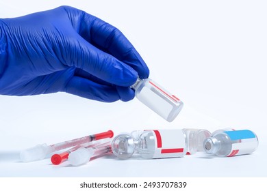 Vaccine vials and needles lie on a white background, a hand in a blue glove holding the vaccine vials above it - Powered by Shutterstock