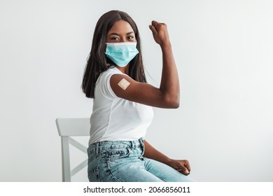 Vaccines Save Lives. Happy Black Lady In Mask Showing Arm With Adhesive Bandage After Covid Antiviral Shot, Vaccinated Young Female Demonstrating Biceps With Plaster, Isolated White Studio Background