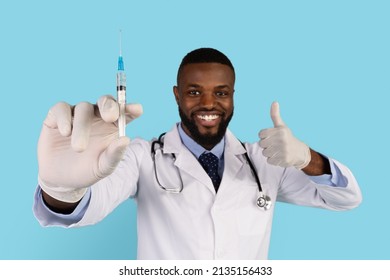 Vacination Campaign. Portrait Of Smiling Handsome Black Doctor With Syringe In Hand Looking At Camera And Showing Thumb Up, African American Therapist In Uniform Posing Over Blue Background