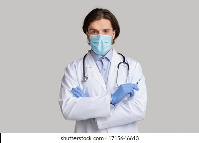 Vacination Concept. Confident Male Doctor In Protective Medical Mask Holding Syringe With Vaccine And Looking At Camera, Light Studio Background