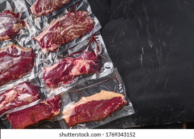 Vacuum packed organic raw beef alternative cuts: top blade, rump, picanha, chuck roll steaks, over black textured background, top view space for text.