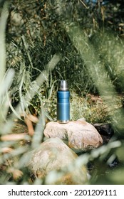 Vacuum Thermos, Camping Flask On A Stone By A Stream In The Forest On A Sunny Day. Equipment For Hiking, Camping.