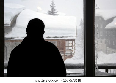 Val D'Isere, Savoi / France - January 17 2018: Silhouette Of A Young Man Looking Out From Window To A Heavy Snow Storm At French Alps. Val D'Isere, Savoie Of France. Buildings Covered With Snow.