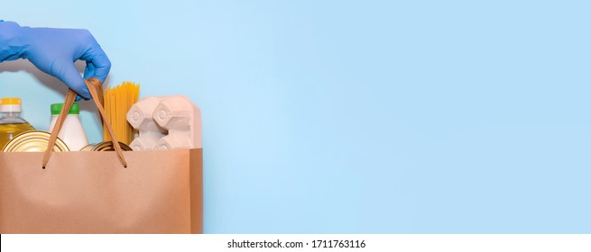 Valanter's Hand In Protective Gloves Gives A Grocery Bag On A Blue Background.Donation Concept. Voluntary Assistance To Those In Need. Copy Space On White Background