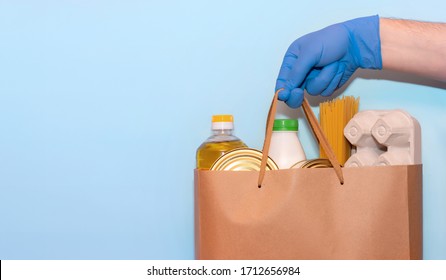 Valanter's Hand In Protective Gloves Gives A Grocery Bag On A Blue Background.Donation Concept. Voluntary Assistance To Those In Need. ?opy Space On White Background