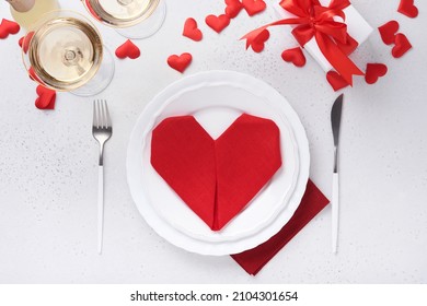 Valentinas day table setting white color with white silverware, red napkin folded as heart, candles, champagne, glasses on white background for greetings. Romantic dinner or menu concept. Mock up.
