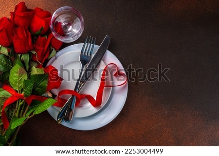 Valentine day table setting on dark background. Romantic diner table setting for with red roses, heart shaped bow, plate, wine glass and cutlery top view copy space. Valentines menu background