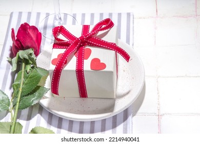  Valentine Day Table Setting With Plate, Gift Box With Festive Red Ribbon, Wine Glass, Fork And Knife, Red Roses Flowers Bouquet White Tiled Table Flatlay Copy Space