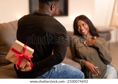 Valentine Gift. Black Boyfriend Congratulating Girlfriend On St. Valentine's Day Holding Wrapped Present Box Behind Back Celebrating Romantic Holiday Together At Home. Cropped, Selective Focus