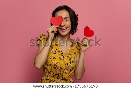 Valentine's cards. Close-up photo of a young charming woman in a floral dress, who is looking in the camera, while covering her eye with one of two heart-shaped cards in her hands.