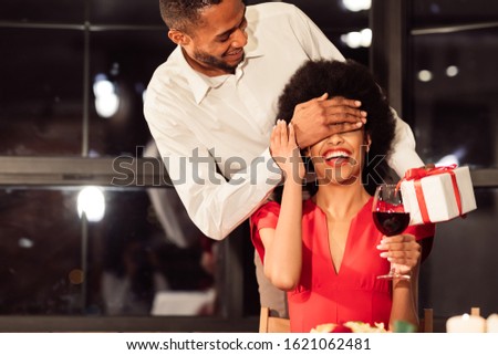 Valentine's Date. Black Man Covering Girfriend's Eyes Giving Gift During Romantic Dinner In Restaurant.