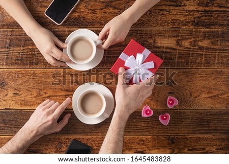 Valentine's Day celebration concept. A nice gift for your loved one. Hands of man and woman with coffee mugs on a wooden table background. Copy space. Flat lay.