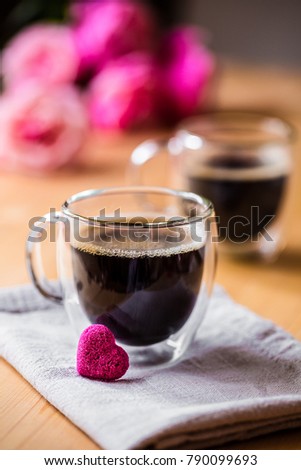 Valentine`s day celebration concept. Two cups of coffee with froth on top on beige napkin in double bottom glasses with heart love shape sugar on wooden table with pink flowers, selective focus