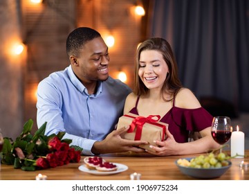 Valentine's Day Celebration. Happy Romantic Interracial Couple Dining In Restaurant, Exchanging Gifts, Loving Black Man Giving Wrapped Present Box To His White Girlfriend, Greeting With Anniversary