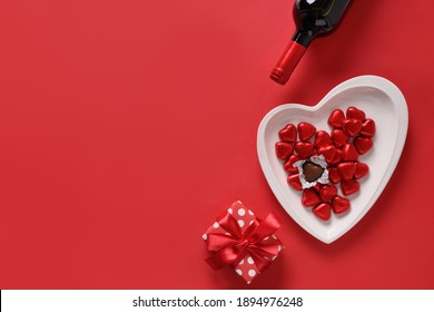 Valentine's day composition of gift, red wine, chocolate sweets on red background. Greeting card with copy space.