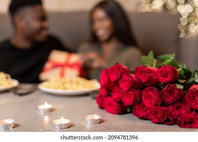 Valentine's Day Concept. African American Couple Celebrating Valentine Holiday Holding Gift Box Sitting At Table With Food And Candles Indoor. Selective Focus On Bouquet Of Red Roses. Shallow Depth