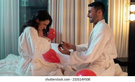 Valentines Day Concept. Beautiful Ethnic Couple Sitting On The Bed Of A Fancy Hotel Room As The Handsome Latino Man Asks Her To Marry Him, Woman Looking At The Ring In Shock, Romantic Theme.