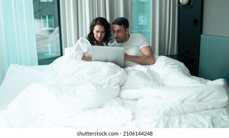 Valentines Day Concept. Beautiful Ethnic Couple Laying On The Bed Under A Blanket In A Fancy Hotel Suite Watching A Movie On A Laptop Device As The Celebrate Valentines Day, Spending Time Together.