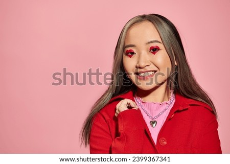 Valentines day concept, cheerful asian woman with heart shaped eye makeup smiling on pink backdrop