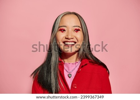 Valentines day concept, cheerful asian woman with heart shaped eye makeup posing on pink backdrop