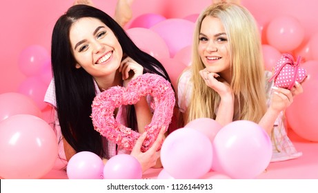 Valentines day concept. Girls lay near balloons, holds heart toys, pink background. Blonde and brunette on smiling faces dreaming about love and date. Sisters, friends in pajamas at pajamas party.
