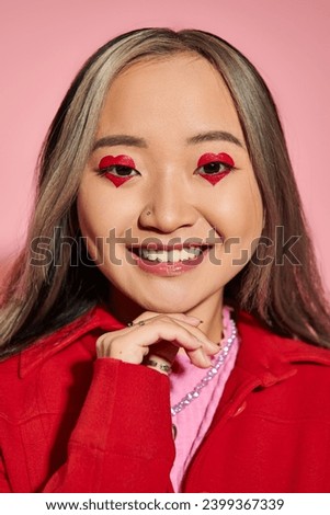 Valentines day concept, happy asian woman with heart shaped eye makeup smiling on pink backdrop
