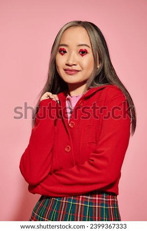 Valentines day concept, jolly asian woman with heart shaped eye makeup smiling on pink backdrop