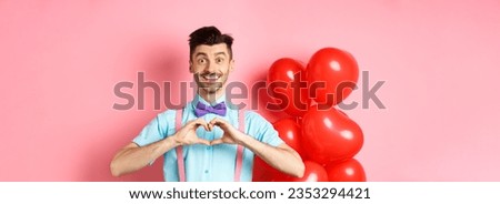 Valentines day concept. Romantic guy in bow-tie, showing heart gesture and say I love you with hopeful smile, standing on pink background near balloons.