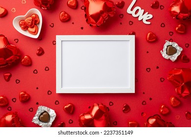 Valentine's Day concept. Top view photo of white photo frame heart shaped balloons chocolate candies plate inscription love and confetti on isolated red background with copyspace