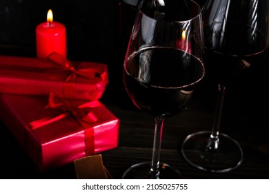 Valentine's Day Concept, Wrapped Gift Box With Red Ribbon, Red Wine Glass, With Red Candle, Piecec Of Chocolate On Dark Wooden Background