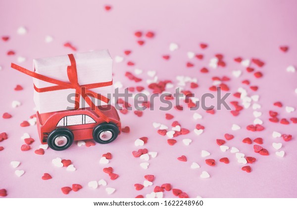 Valentines day delivery concept. Miniature red car\
with a present box on the roof on a pink background with red and\
wihte hearts. Toning\
image