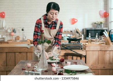 Valentines day dinner with table place setting up indoor alone prepared process. flower in vase next to champagne glasses on wooden kitchen table. young girl putting red rose on white plate. - Shutterstock ID 1334975846