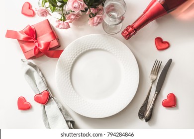 Valentines day dinner with table place setting with red gift, glass for champagne, a bottle of champagne, pink roses, hearts with silverware on white background. View from above. Valentine's card.