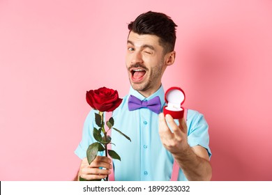 Valentines day. Funny guy making proposal, winking and saying marry me, showing engagement ring with red rose, standing over pink background
