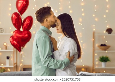 Valentines day. Handsome young man kissing woman forehead while she leaning into him. Happy couple hugging each enjoying tender romantic moment against background of red heart shaped balloons - Powered by Shutterstock