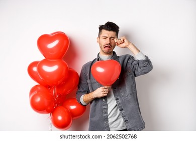 Valentines day and love concept. Sad crying man holding red heart balloon and whiping tears, standing single and miserable, being heartbroken, white background