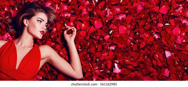 Valentine's Day. Loving girl. The girl in a red dress lying on the floor in the petals of red roses. Background of red rose petals. Red lipstick on the lips from the beautiful girl.