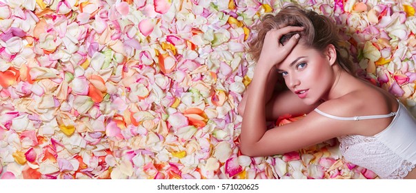 Valentine's Day. Loving girl. The girl in white dress lying on the floor in the petals of roses. Background of white, orange, red, pink rose petals. Pink lipstick on the lips from the beautiful girl.