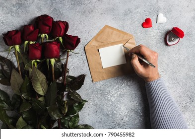 Valentines Day Man Hand Holding Pen Writing Love Letter With Greeting Card Mothers Day Red Rose Gift Surprise On Grey Background With Copyspace. Love Flower Gift For Woman Romantic Holiday Birthday