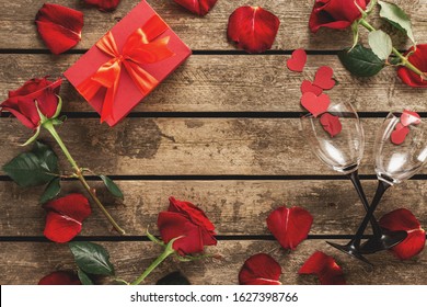 Valentine's Day Or Mother's Day Concept. Frame Of Red Gift Box With Flowers, Rose Petals And Wine Glasses On Wooden Background Top View