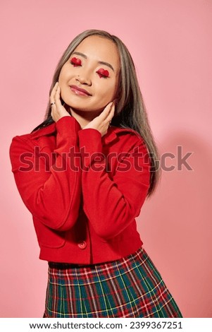 Valentines day, pleased asian young woman with heart eye makeup posing in red jacket on pink