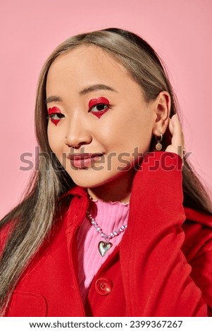 Valentines day, positive asian young woman with heart eye makeup posing in red jacket on pink