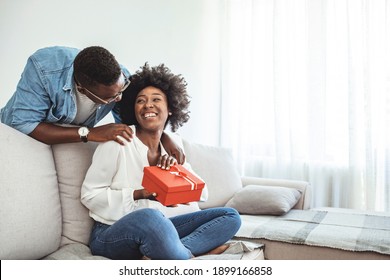 Valentines Day, Relationships And People Concept - Happy Couple With Gift Box Hugging At Home. Young Man Presenting Gift To His Girlfriend. Valentine's Day Celebration