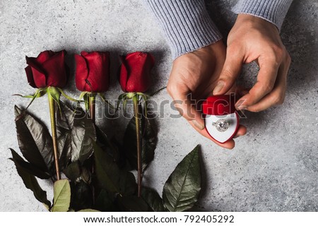 Valentines day romantic man hand holding engagement ring in box marry me wedding with red roses bouquet gift surprise on grey background. Love gift for woman making proposal
