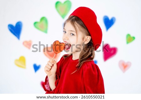 valentine's day, a small smiling child girl in a red beret happy holds or licks a large lollipop in the form of a heart and rejoices against the background of colorful hearts.