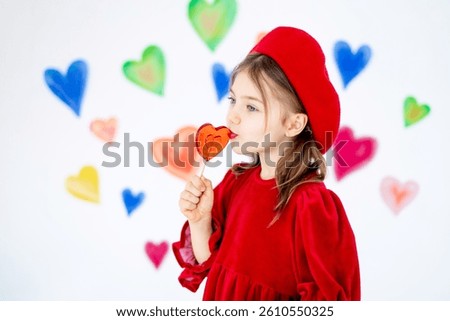 valentine's day, a small smiling child girl in a red beret happy holds or licks a large lollipop in the form of a heart and rejoices against the background of colorful hearts.