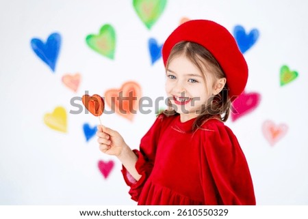 valentine's day, a small smiling child girl in a red beret happy holds or licks a large lollipop in the form of a heart and rejoices against the background of colorful hearts.