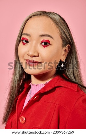 Valentines day, smiling asian young woman with heart eye makeup posing in red jacket on pink