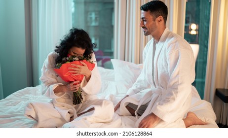 Valentines Day Special. Beautiful Young Ethnic Couple Sitting On The Bed Of A Fancy Hotel Room. Woman Holding A Cute Flower Bouquet That Her Boyfriend Gifted Her, Looking Extremely Happy.